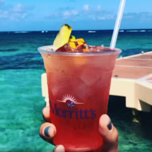 A person's hand holding a right pink and red cocktail against a clear blue sea 